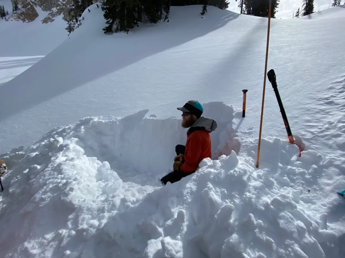 A skier in a red jacket standing in a snow pit with a probe pole beside it