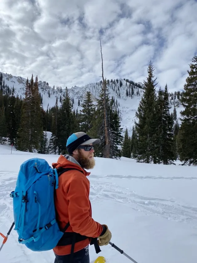 Skier in an orange jacket and blue backpack on a snowy slope with mountains behind