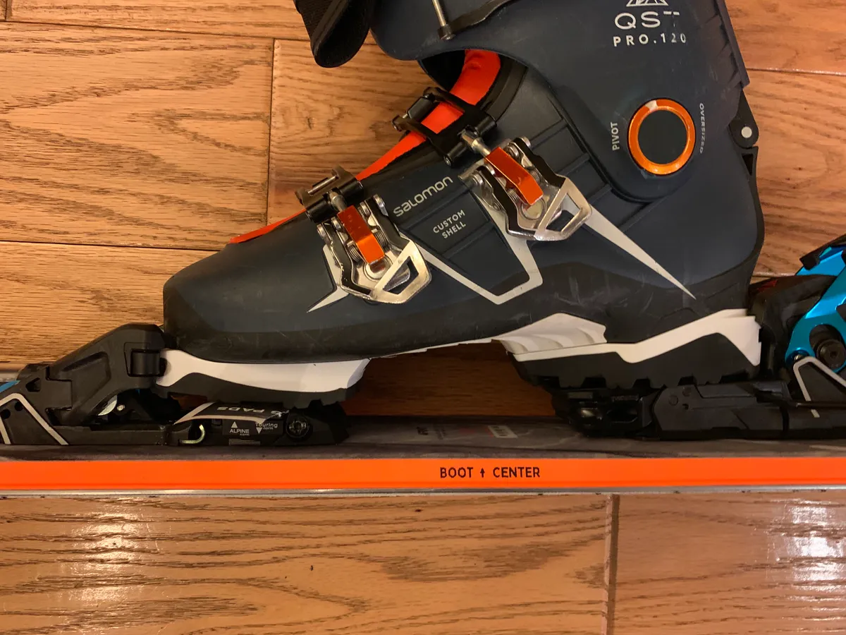 Salomon ski boot clipped into a binding on an orange ski on a wood floor